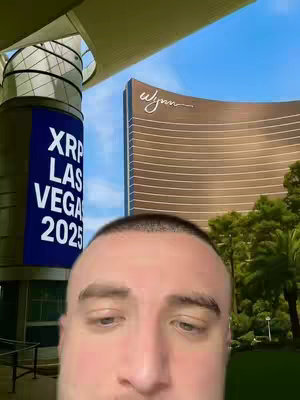 A selfie with a man in the foreground, the background features a large building with the Wynn hotel logo, and a blue sign that says "XRP LAS VEGAS 2025". The sky is a clear blue.