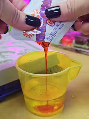 A close-up shot shows a gloved hand pouring a stream of orange liquid from a white and red packet into a clear yellow plastic cup. The cup is partially filled with a greenish granular substance, and a layer of the orange liquid has settled at the bottom. The background includes soft, blurred colors, and various items out of focus.