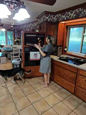 A woman stands in a warm-toned kitchen, holding a spatula and looking into a built-in microwave. She's wearing a gray shirt and a checkered skirt, and her hair is pulled back. A dog is walking in the foreground, and the kitchen features wooden cabinets, a tiled floor, and a window. The overall setting suggests a home environment.