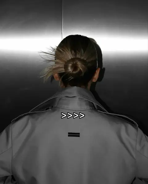 The cover image shows a person from the back, wearing a gray trench coat, facing a metallic, reflective surface. The person's hair is styled in a bun. Text overlays the image at the top and bottom. The person could be a girl.