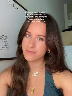 A woman with long brown hair and blue eyes is facing the camera with a slight smile. She is wearing a teal top and two gold necklaces with pendants. Behind her is a whiteboard and a wall.