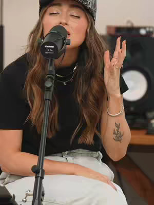 A woman with long brown wavy hair is singing into a microphone. She's wearing a black t-shirt, light colored pants, and a patterned baseball cap. She has a tattoo on her forearm and is wearing jewelry. The background is blurred, with studio equipment visible.