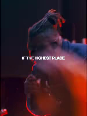 The image is a close-up shot of a person from the shoulders up, possibly a musician performing. They have dark hair with what appears to be dreadlocks. The person is holding something, possibly a microphone, with their right hand, partially obscured by a red-orange light. The setting has a moody, dark atmosphere with red and black colors dominating the backdrop. Text overlayed reads, 'IF THE HIGHEST PLACE' in white.
