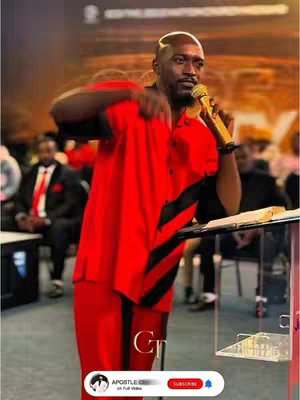 A man in a red suit with black stripes on the side is holding a microphone and speaking in front of a podium. The image is well-lit with a blurred background of an audience.