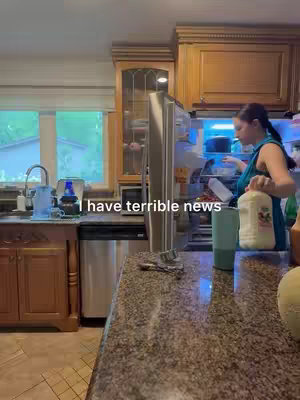 A woman stands in a well-lit kitchen, holding a container of milk near an open refrigerator. The background reveals wooden cabinetry and a large window illuminating the domestic setting. Text overlayed on the image reads, "I have terrible news."