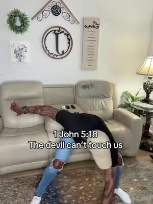 A man is bent over backward, forming an inverted V-shape, with his hands on the floor and legs bent at the knees. He is wearing a black t-shirt and ripped blue jeans. The background is a room with a white sofa, a lamp, and wall decor. Text overlays read '1 John 5:18' and 'The devil can't touch us'.