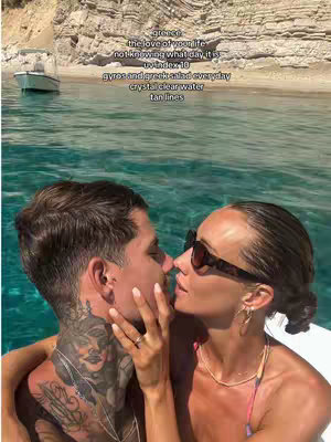 A couple is seen close together, about to kiss. The male has a neck tattoo, while the woman has sunglasses on. Behind them is clear blue water and a sandy beach with a boat.