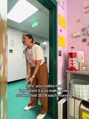 A woman in a classroom doorway is leaning forward with a smile. Text overlayed on the image says 'POV: you realize how important it is to make every student feel SEEN each morning'. The classroom's interior is mostly pink with decorative elements, a red gumball machine, and shelves with storage bins.