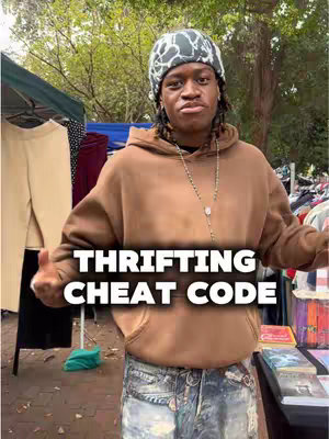 A young black man stands in front of a clothing rack in an outdoor market setting. He wears a beanie, necklace, and hoodie, and has a text overlay reading 'Thrifting Cheat Code'.