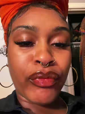 A close-up shot of a person's face features prominently. The person has brown skin, with their eyes closed, and their lips are glossed and slightly parted. They are wearing large hoop earrings and a head covering. The composition is tightly framed, focusing only on the face and upper neck.