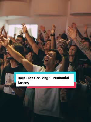 A crowded room filled with people with their arms raised in worship. The focus is on a man in a white t-shirt with his hands lifted towards the ceiling. He's singing with his eyes closed, surrounded by others with similar expressions. The lighting is warm, suggesting a spiritual or religious gathering.