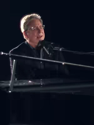 A man with glasses is singing and playing the piano. The background is dark. The lighting suggests a stage setting.
