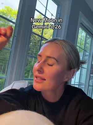 A woman with blonde hair and an updo is looking slightly upwards, with a window and greenery in the background. Text overlaid on the image reads, "New Moon in Gemini 5/26".