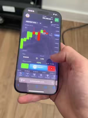 A person's hand holding a smartphone displaying a trading platform interface. The screen shows a graph with green and red candlesticks, indicating market fluctuations. Various buttons and data points, including the account balance and trading options, are visible. The background is neutral, with focus on the phone and the user's hand.