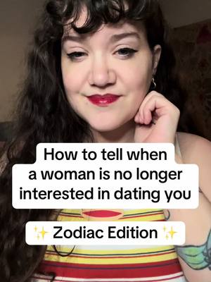 A woman with long, wavy dark hair is shown in a close-up shot, angled slightly down. She has fair skin, full red lips, and dark eyeliner. Her left hand is gently touching her cheek. She's wearing a striped top with a yellow, white, and red pattern. Text overlays in a white box read: 'How to tell when a woman is no longer interested in dating you Zodiac Edition.' The background is slightly blurred, appearing to be a room.