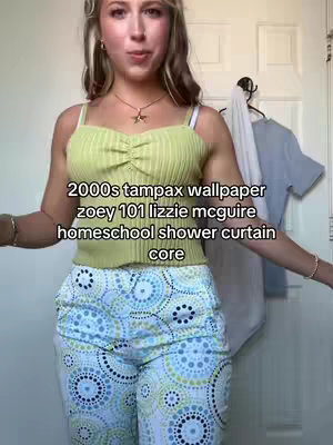 A woman stands in front of a neutral background. She is wearing a yellow-green ribbed tank top and patterned blue and white capri pants. The background is slightly out of focus, but a glimpse of a door and a shirt hanging from a doorknob is visible.