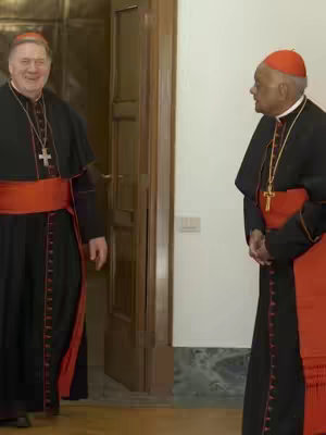 Two cardinals in ceremonial attire stand in a well-lit hallway. The cardinal on the left stands near an ajar wooden door, gazing towards the other figure. The second cardinal stands further down the hallway, facing the first and slightly to the left, hands clasped in front. Both figures are positioned against a plain white wall and the door. The lighting suggests an interior setting, with natural or diffused light.