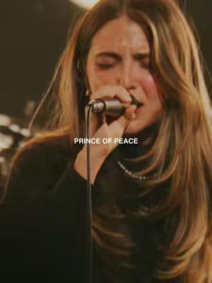 A medium shot of a woman singing into a microphone. Her face is partially obscured by the microphone, but her eyes are closed as she sings. The background appears to be a performance venue, and the lighting is warm. Text overlayed on the image reads 'PRINCE OF PEACE'.