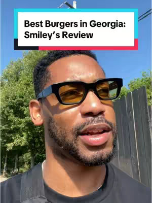 A selfie-style video cover image featuring a man with short black hair, a well-groomed beard, and dark-framed sunglasses with yellow-tinted lenses. The man is looking slightly off-camera with a neutral expression. Above him, a brightly colored banner reads "Best Burgers in Georgia: Smiley's Review". The background is blurred, suggesting an outdoor setting with hints of greenery and a clear, sunny sky.