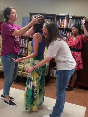 The image depicts four women indoors, likely in a home or church setting. One woman in a maroon shirt and another in a white shirt support the middle woman, who wears a blue tank top and patterned skirt. The woman in the maroon shirt has her arms raised with expression. Bookshelves and furniture are visible in the background. The color palette is warm, with natural light.