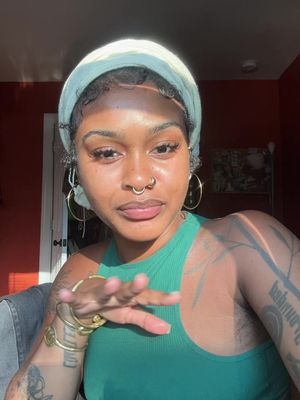 A selfie of a woman with dark skin, wearing a green tank top and light green head wrap. She has several gold accessories including hoop earrings, nose ring, and bracelets. She is holding out her hand toward the camera, with tattoos visible on her left arm. The background is a warm-toned wall with a window showing sunlight.