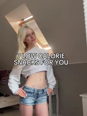 A woman in a white crop top and denim shorts stands in a room with a sloped ceiling. Text overlayed reads \"7 LOW CALORIE SNACKS FOR YOU\".