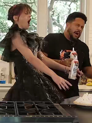 A woman in a formal black dress and a man in a black t-shirt are covered in cream, mid-explosion from what appears to be a cream pie. They are in a kitchen, with the woman's mouth open wide and the man holding two cans of whipped cream. A pie is on the counter. The background has white cabinets and windows with natural light.