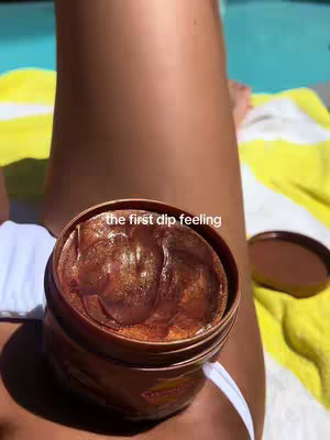 An overhead shot shows a leg with bronzed skin, a tanning product container, and a yellow towel by a pool. The product is in an open dark brown jar. Above the jar, text reads 'the first dip feeling'.
