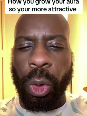 The cover image is a close-up shot of a Black man's face. The man has a bald head and a dark, neatly trimmed beard. His eyes are closed, and his lips are slightly parted, as if he is exhaling. The background is a blurred, warm-toned interior. In the top area of the image, the text overlay reads, "How you grow your aura so your more attractive."