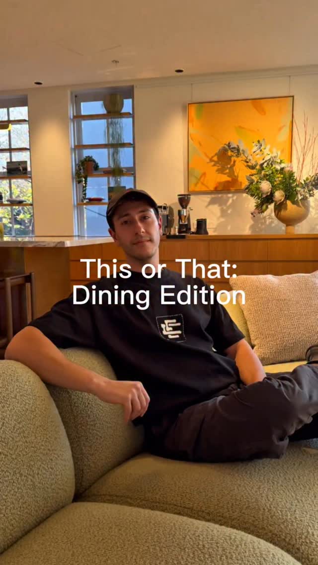 A man wearing a black shirt and a baseball cap is seated on a green couch. The text overlay says "This or That: Dining Edition". The background includes a window, artwork, and a small cabinet.