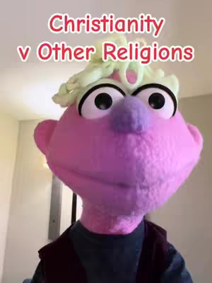 The cover image features a puppet with large eyes and a fuzzy, pink face wearing a blue shirt and a maroon jacket. The puppet is wearing a crown-like accessory and the white background is very neutral. Text overlay in the top-center of the image reads 'Christianity v Other Religions'.