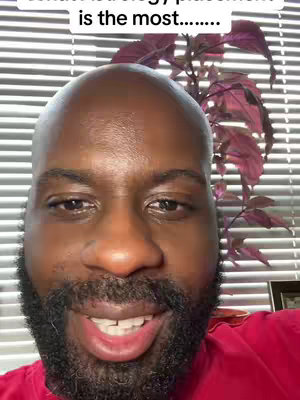A close-up shot of a Black man's face, with a shaved head and full, dark beard, smiling slightly at the camera. The background includes horizontal blinds and a plant with burgundy leaves.