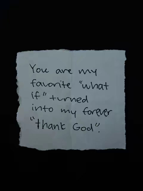 A close-up photograph shows a small, torn piece of off-white paper with handwritten text against a stark black background. The paper has rough, uneven edges, suggesting it has been ripped. The handwritten text, in a simple, slightly irregular font, reads: 'You are my favorite "what if" turned into my forever "thank God".' The black backdrop enhances the contrast, making the text stand out and adding a sense of intimacy and personal touch, likely due to handwriting. The image is well-lit, showing the paper's texture.