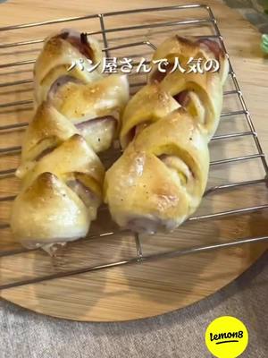 The cover image displays two braided bacon-wrapped pastries placed on a metal cooling rack set atop a wooden cutting board. The pastries are golden brown with visible textures and a hint of purple fillings peeking through. The background features a lightly blurred, neutral surface, with a yellow logo in the bottom right corner.