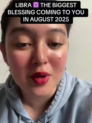 A medium shot featuring a woman looking at the camera. She has fair skin, dark hair pulled back, and is wearing a light blue hoodie. The background is plain and white. A text overlay in a black box reads, 'LIBRA ♎️ THE BIGGEST BLESSING COMING TO YOU IN AUGUST 2025'. The woman has red lipstick and visible blush, appearing to be in a well-lit setting.