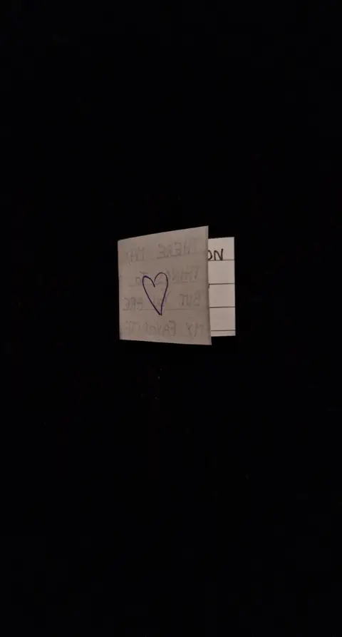 The image shows a piece of paper with a heart drawn on it, with text partially visible. The background is solid black.