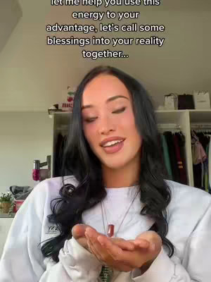 A woman with long dark hair and a white sweater is centered, facing slightly towards the side with closed eyes. She is holding her open palms towards the viewer. Above her, a black text overlay states 'Let me help you use this energy to your advantage, let's call some blessings into your reality together...' The background is slightly blurred, showing a closet with clothes and items. The lighting is soft, with a warm overall tone.