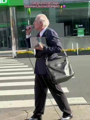 An older man, wearing a dark blue suit, white shirt and red tie, is walking on a crosswalk while holding a microphone in one hand and a small clear book in the other. He is speaking into the mic. A black bag hangs over his shoulder. The background shows a city street with modern buildings.