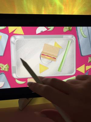 The image shows a person's hand with a stylus interacting with a tablet or computer screen, displaying a game interface. The interface has a background featuring pink and a digital tray with a sandwich, cheese triangles, and a celery stalk. The foreground includes the hand and stylus, positioned towards the sandwich on the tray. To the left are broccoli and other design elements arranged in a circular fashion.