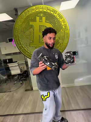 A man stands in an office setting, holding objects in both hands. Behind him is a large, golden bitcoin symbol. In the background, translucent panels and office furniture are visible.