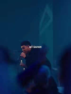 A person, likely a singer, is holding a microphone and singing passionately. The background is a blurred blue with stage lighting. The text overlay says: MY SAVIOR.