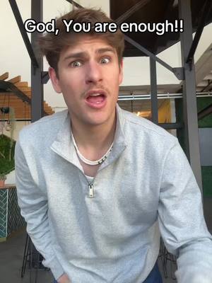 A medium shot features a young man with short, light brown hair and a surprised expression. His eyes are wide, and his mouth is open as if he's speaking. He is wearing a light gray, zipped-up sweater, over a white shirt and a beaded necklace. The background includes metal structures and glimpses of an outdoor setting.