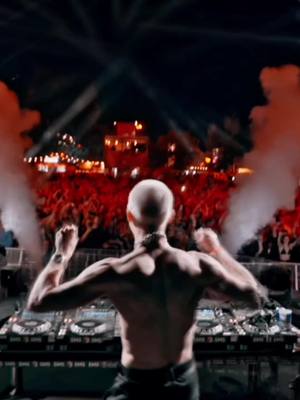 A shirtless man with his back to the camera stands behind a DJ setup in front of a crowd at a concert. The stage is lit with bright lights, and smoke fills the air.