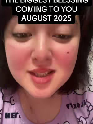 A close-up shot features a light-skinned woman with fair skin and blushed cheeks. She’s smiling slightly and wearing a purple top with white patterns. Overlaying the image, a black banner with white text reads 'THE BIGGEST BLESSING COMING TO YOU AUGUST 2025'.