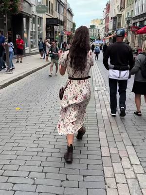 A street scene, viewed from behind a woman walking directly away from the camera. The woman is wearing a long floral dress and boots. The cobblestone street is lined with buildings and pedestrians, and the overall tone is natural and documentary-style.