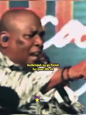 The image shows a man with a bald head and a dark complexion, possibly of African descent. He appears to be singing or speaking with great emotion, his mouth open and eyes wide. He's wearing a patterned shirt, and a microphone is visible near his face. Text overlays the image, and a logo is present in the lower-left corner. The background is blurred but includes bright colors.