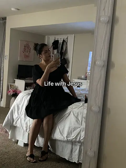A woman in a black dress sits on a bed, taking a selfie with her phone. A large mirror with lights frames the shot. Soft lighting and a bedroom setting provide the background.
