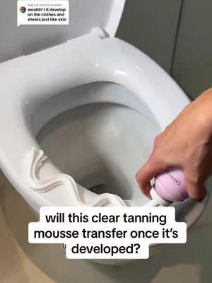 A hand holding a pink and white can of tanning mousse over a white toilet. The toilet lid is up, and there is a curved decorative piece on the front of the bowl. Text at the bottom asks if the mousse will transfer once developed, and a comment in the upper left corner discusses potential transfer to clothes and sheets.