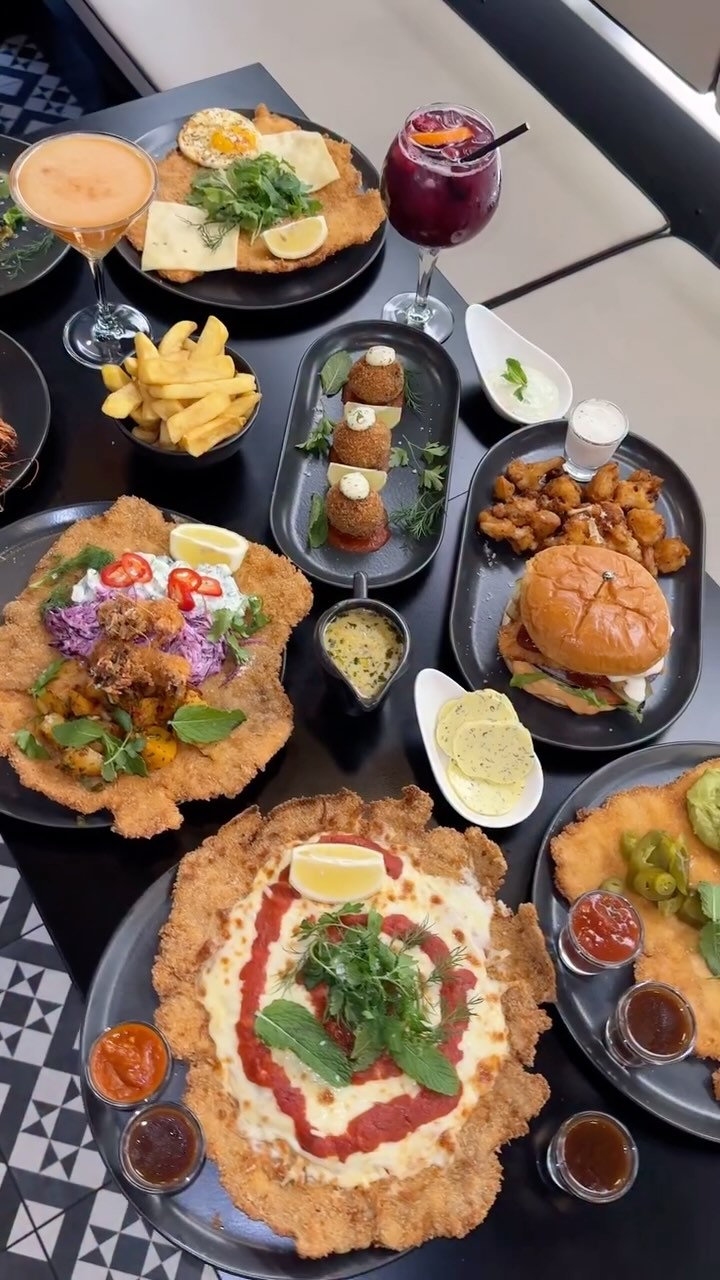 A top-down shot featuring a variety of schnitzel dishes artfully arranged on a dark table. The schnitzels are the main focus, showcased with an array of toppings and sides. The composition is well-lit, highlighting the textures and colors of the food, with plates and small dishes of sauces and garnishes adding visual interest.