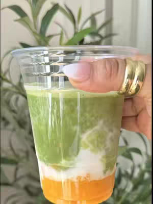 The image shows a layered drink in a clear plastic cup held by a hand with a gold ring. The drink consists of three distinct layers: an orange base, a white middle layer, and a green upper layer. The background shows blurry green leaves.